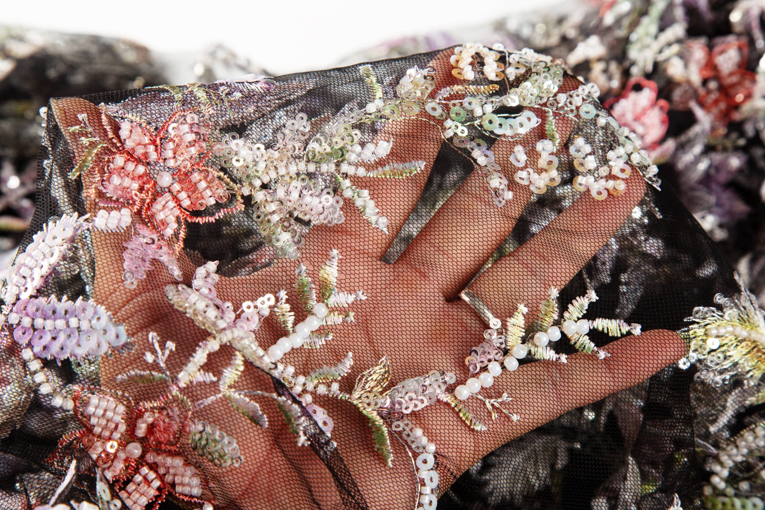 Close-up of sheer black mesh fabric featuring flat floral embroidery seamlessly integrated with vibrant pink and green sequins and glass beads, demonstrating complex multi-craft manufacturing.
